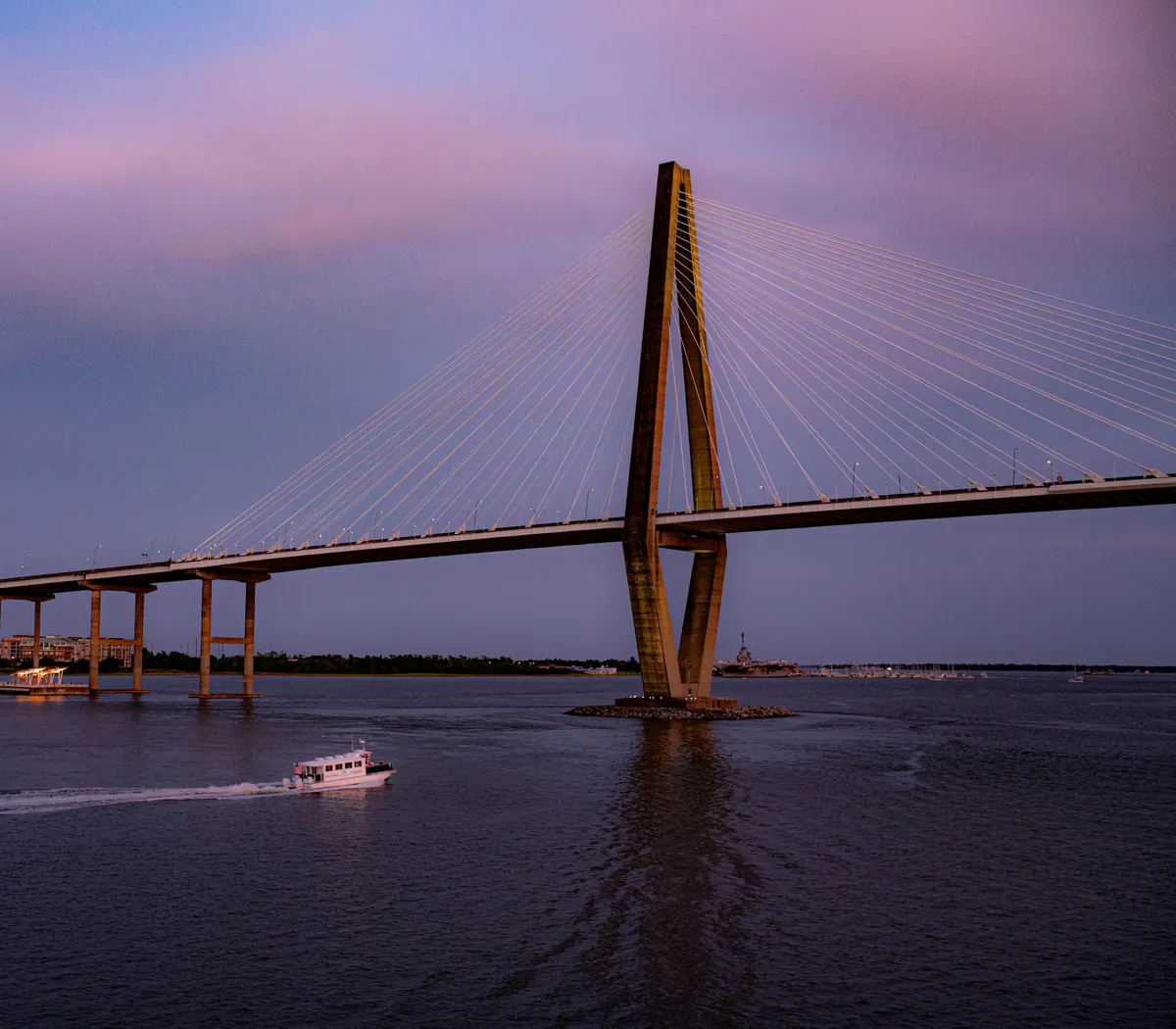 One tower of the Ravenel Bridge and its cables against a violet dusk sky, with a small white boat leaving a wake on the dark water below