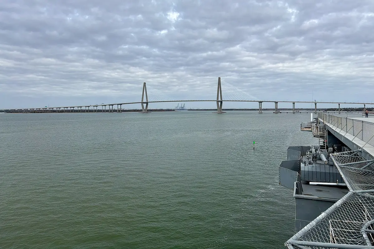 The long, low line of the Ravenel Bridge with its two towers seen across the grey-green water of the harbor from the deck of the USS Yorktown, under a cloudy sky