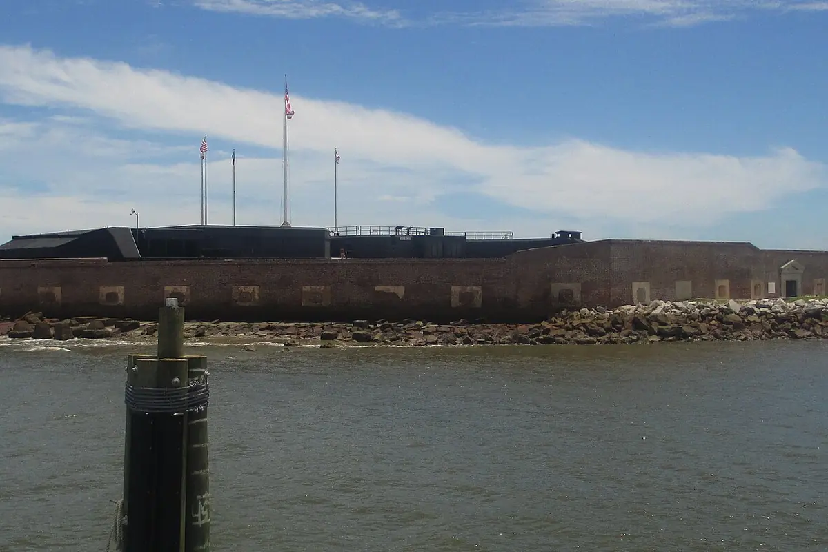 Die niedrigen Ziegelmauern von Fort Sumter auf seiner künstlichen Insel, mit Fahnenmasten über der Mauer und Felsen entlang der Wasserlinie unter blauem Himmel