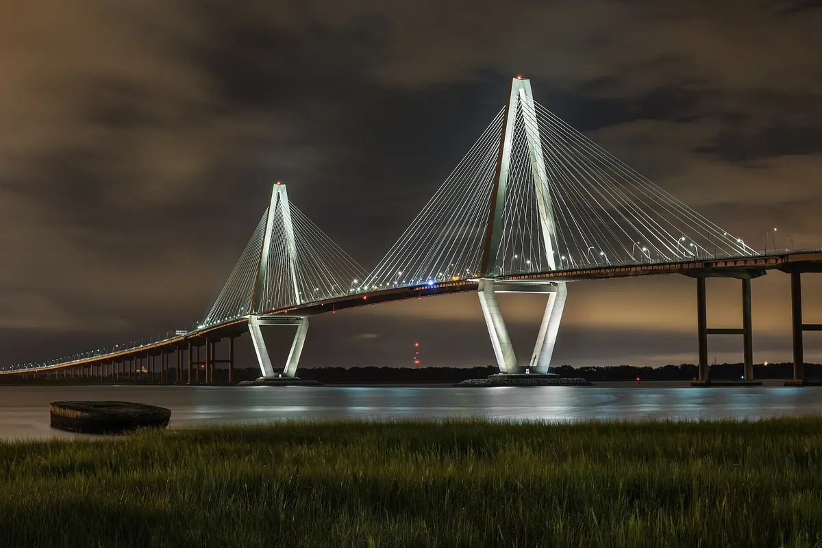 Die Arthur Ravenel Jr. Bridge bei Nacht, ihre beiden diamantförmigen Türme und das Fächerwerk der Kabel spiegeln sich im Cooper River, im Vordergrund Marschgras
