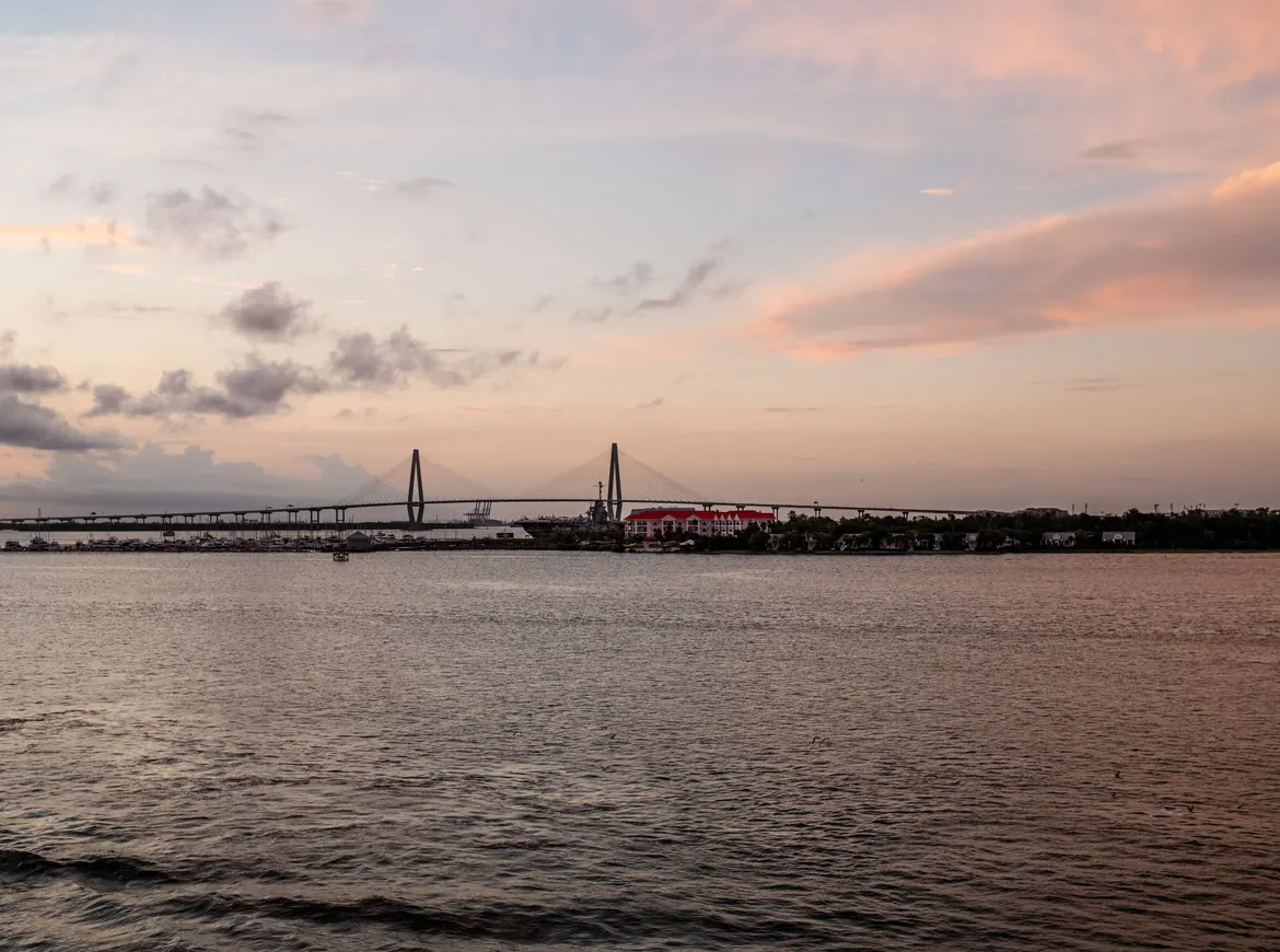 A wide harbor at sunset with the distant Ravenel Bridge towers on the shore, under pink and orange clouds